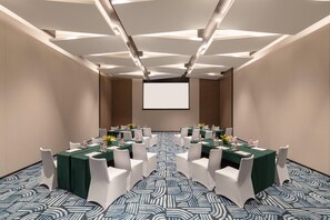 Une salle de conférence dotée d’un grand écran à l’avant, de plusieurs tables entourées de chaises et d’un tapis à motifs.