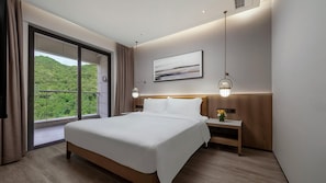 Ein modernes Hotelzimmer mit einem großen Bett, einem Nachttisch mit Lampe, Blick auf Grün durch das Fenster und einem Gemälde an der Wand.