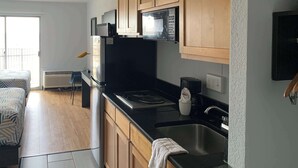 A compact kitchen with wooden cabinets, a black countertop, a built-in microwave, and a sink. - Studio 6 Lincoln, NE - Airport (Lincoln)