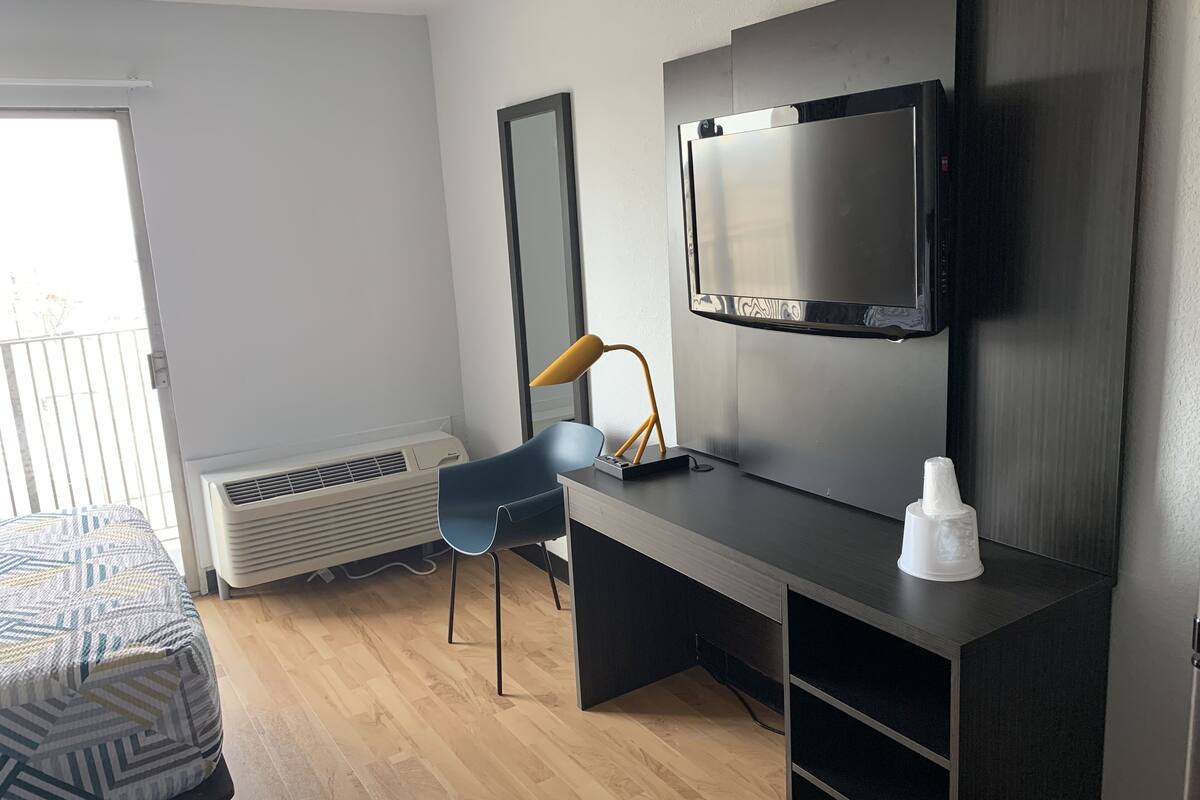 A modern hotel room with a large flat-screen TV mounted on the wall, a desk with a chair, a bedside table with a lamp, and a patterned bed.