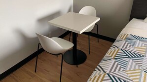 A small, clean room with a white table and two white chairs, a patterned bedspread, and a wooden floor. - Studio 6 Lincoln, NE - Airport (Lincoln)