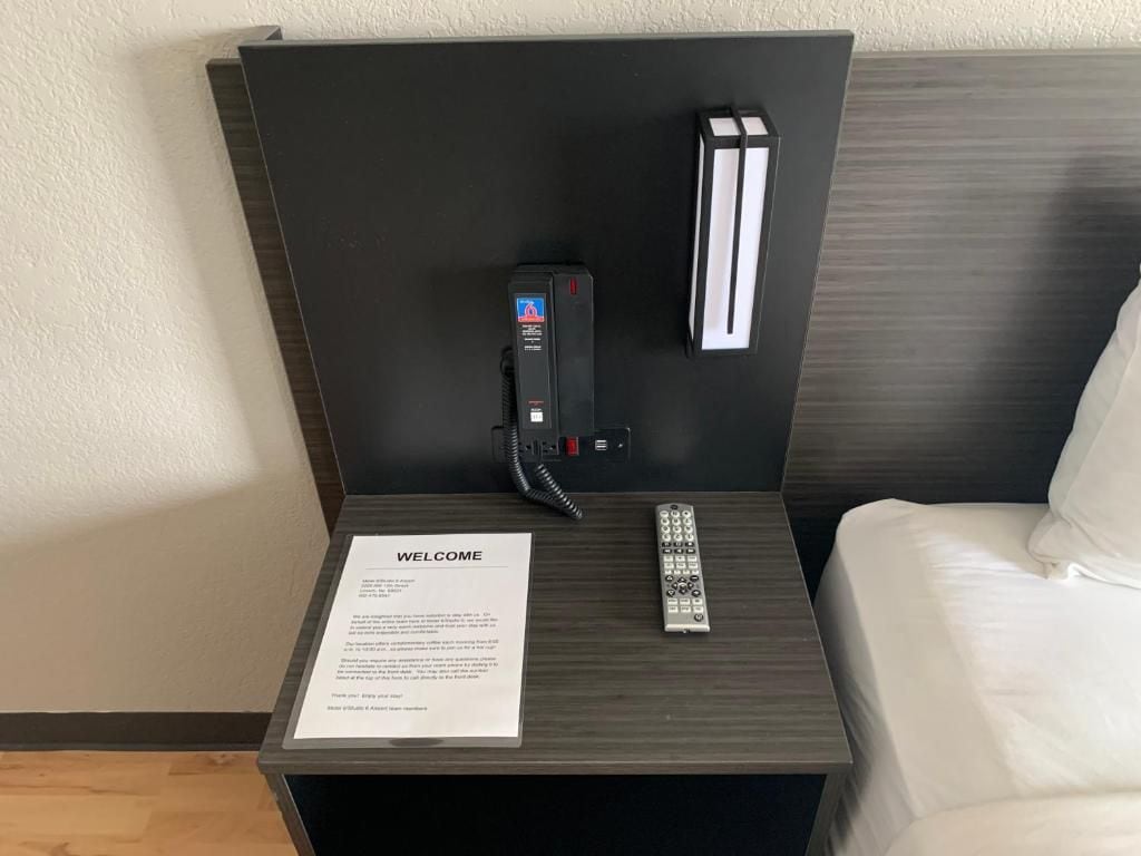 Habitación de hotel con una cama, una mesita de noche con control remoto, un teléfono y un cartel de bienvenida.