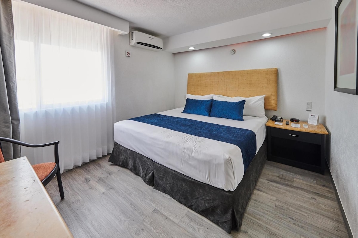 A hotel room with a bed, bedside table, chair, and a window with curtains.