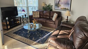 A living room with a leather sofa, a glass coffee table, a TV on a stand, and a large window with curtains.