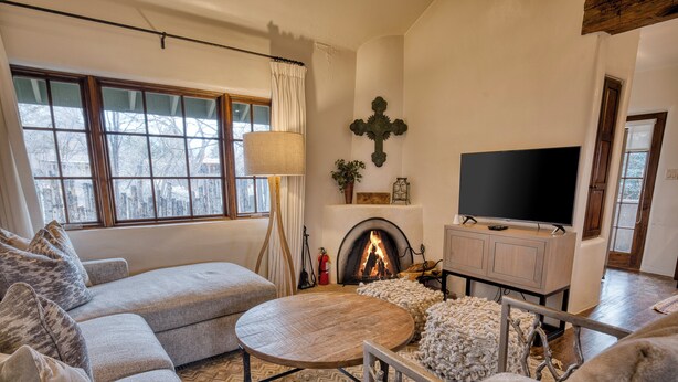 A cozy living room with a fireplace, a television, and a large window with curtains. - Casa Dos Truchas | Adobe Retreat (Santa Fe)