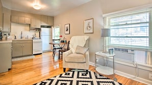 A modern kitchen with wooden flooring, a dining area with a table and chairs, a cozy armchair, and a large window with blinds. - Historic Apartment - Walk to CSU Campus + Old Town (Fort Collins)