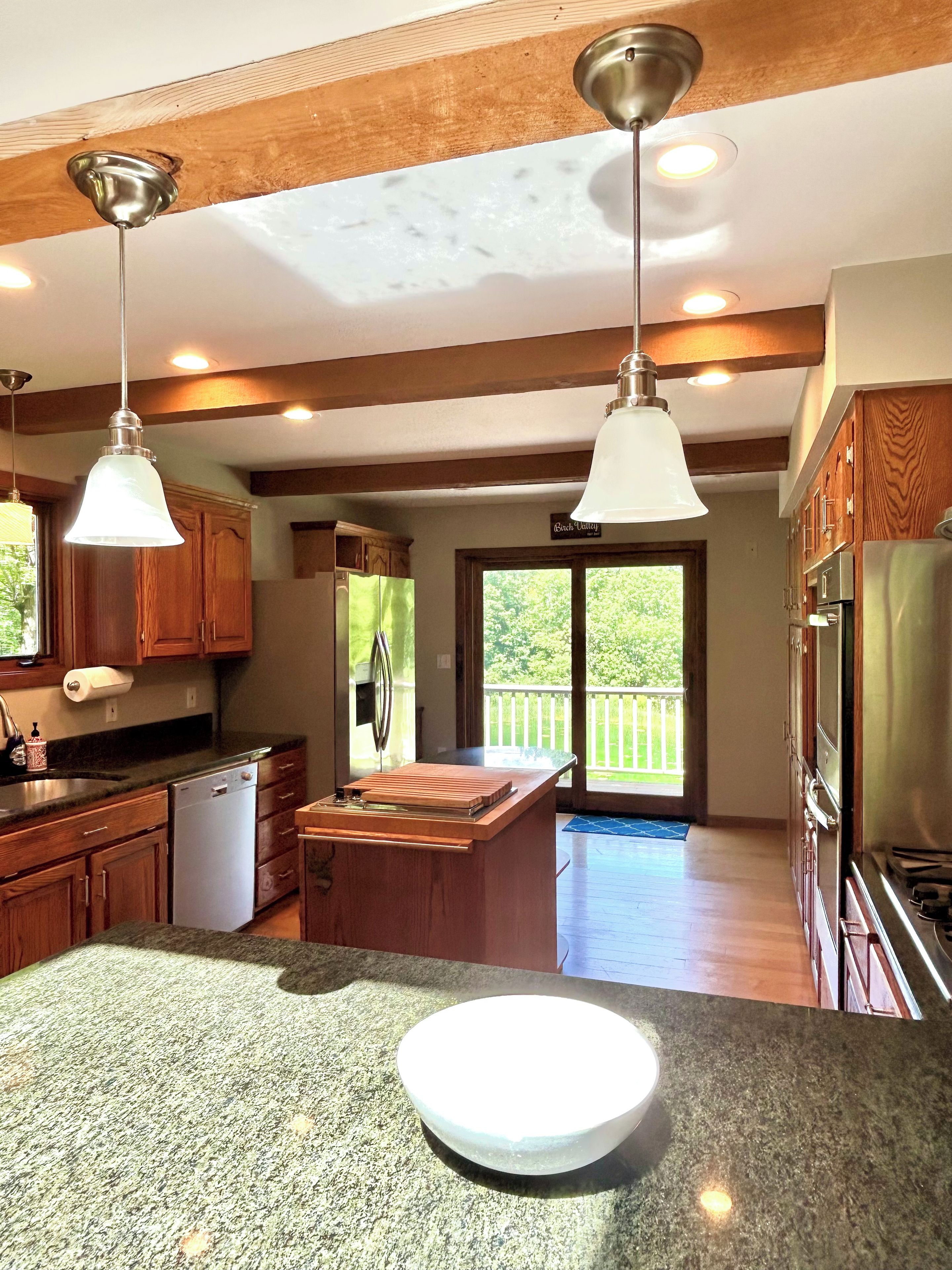 A kitchen with granite countertops, wooden cabinets, stainless steel appliances, pendant lighting, and a view to the outside.