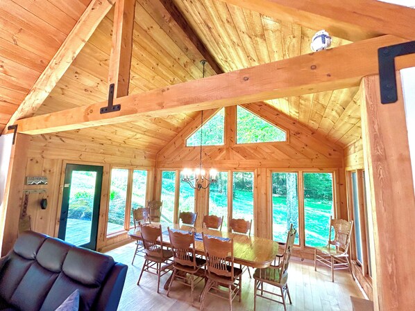A wooden cabin interior with a dining table and chairs, large windows, and a chandelier.