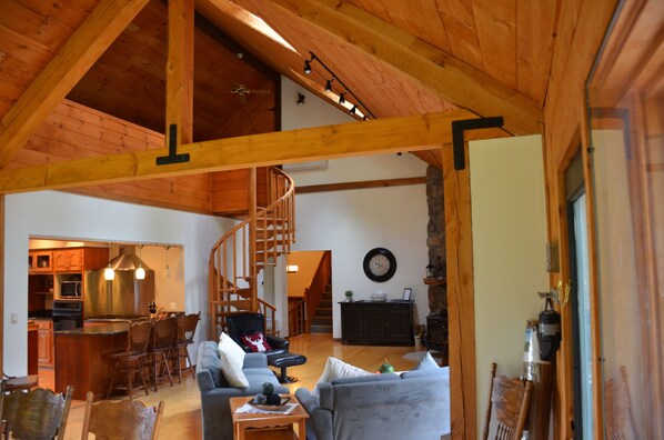 A spacious living area with a high ceiling, wooden beams, a staircase, a kitchen, and a dining area.
