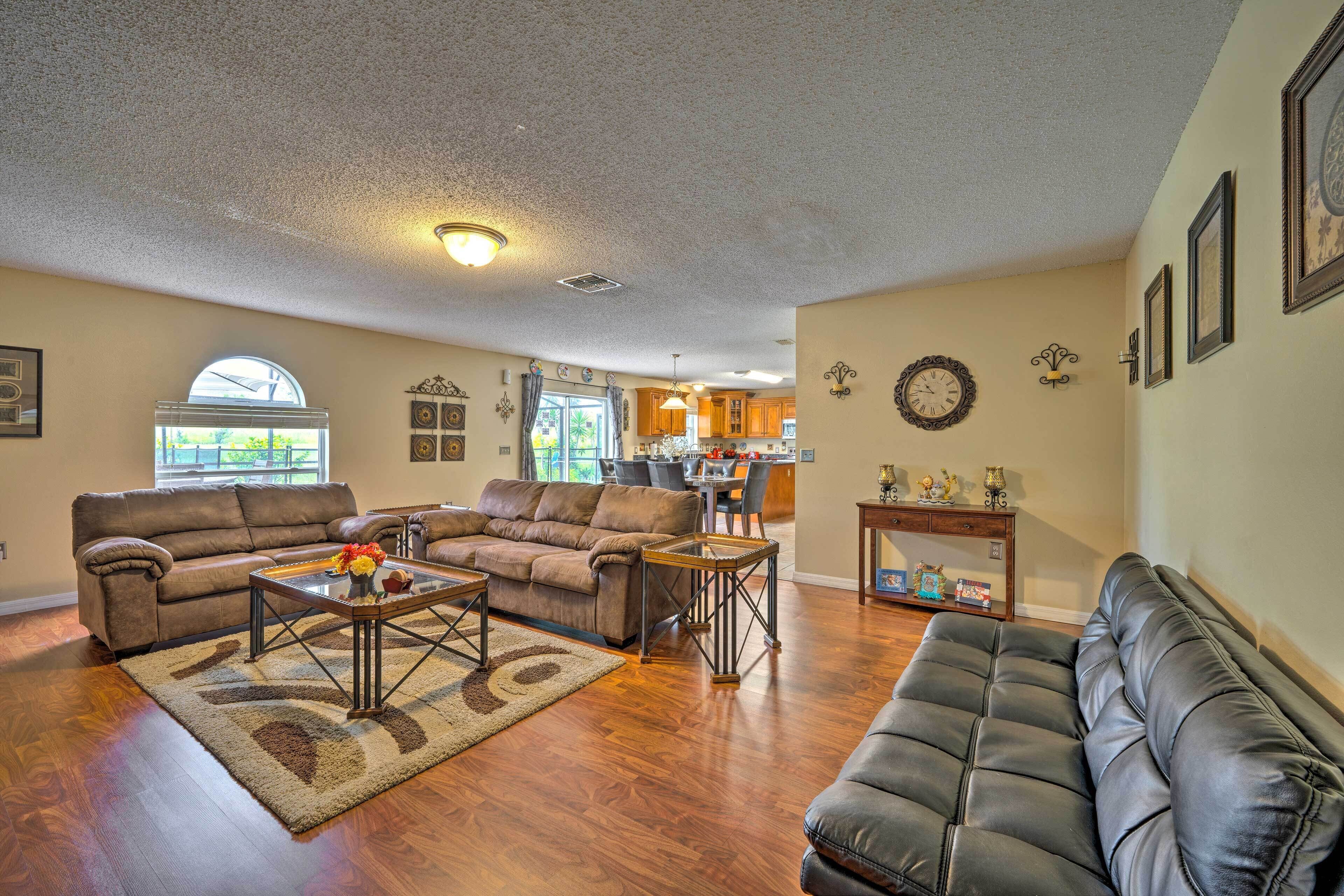 A spacious living room with a brown leather couch, a black leather sofa, and a coffee table. There is a kitchen in the background with wooden cabinets and a dining area.