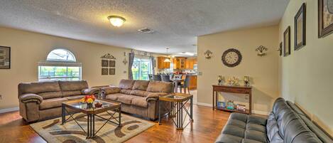 A spacious living room with a brown leather couch, a black leather sofa, and a coffee table. There is a kitchen in the background with wooden cabinets and a dining area.