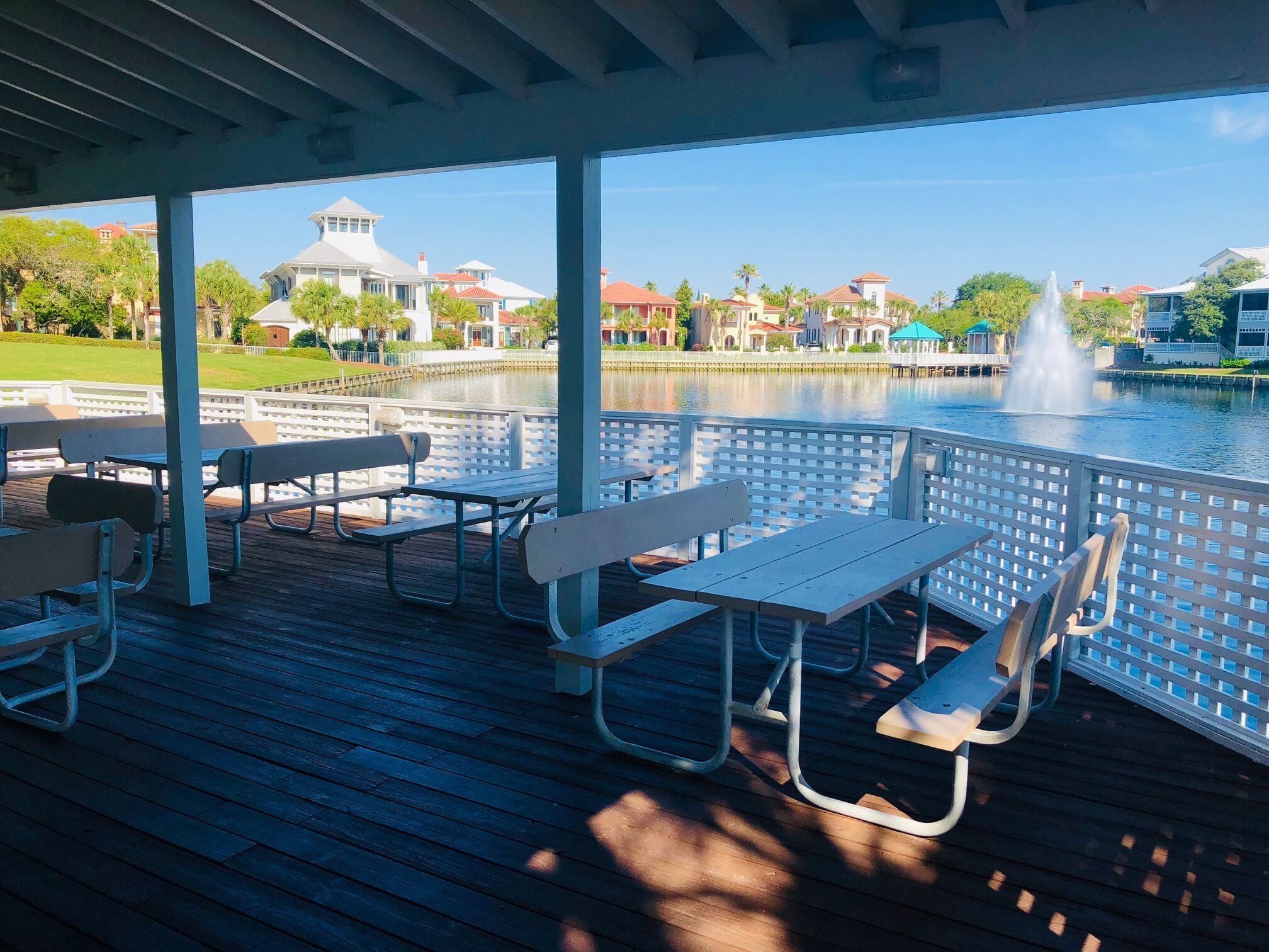 Een terras buiten met picknicktafels en banken, met uitzicht op een waterpartij met een fontein, omgeven door een hek en huizen op de achtergrond.
