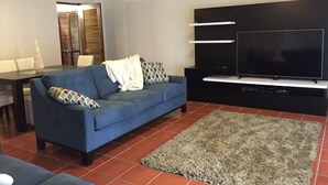 Una sala de estar moderna con un sofá azul, un televisor de pantalla plana y una zona de comedor con mesa y sillas.
