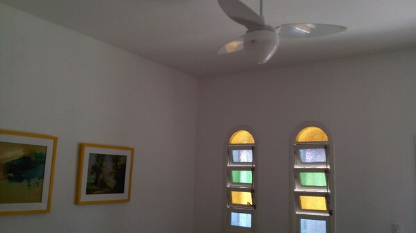 A room with a ceiling fan, two windows with stained glass, and two framed pictures on the wall. - CARAGUATATUBA HOUSE WITH 4 BEDROOMS 5 MINUTES FROM PALMEIRAS BEACH, 5 FROM CIGARRA! (caraguatatuba)