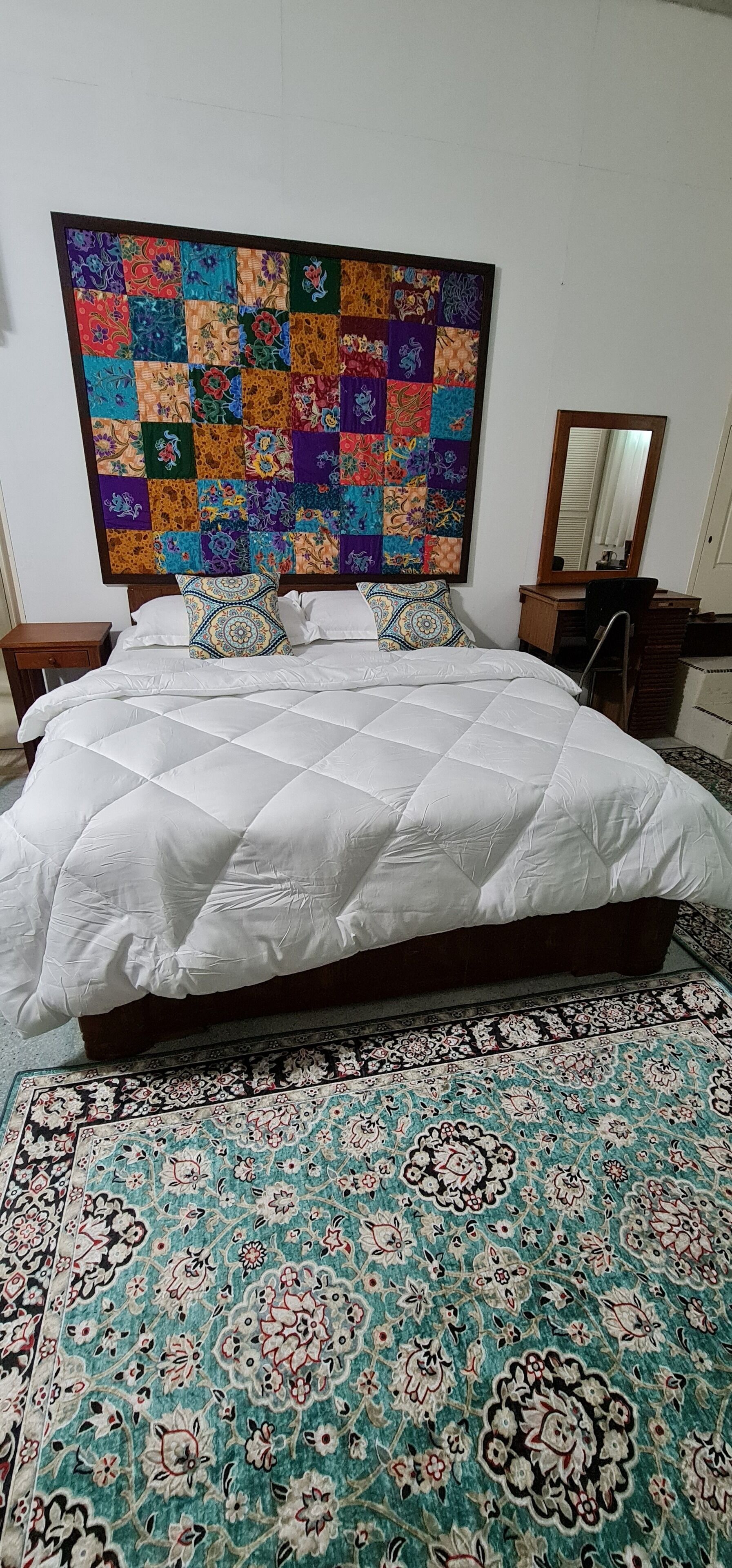 Un lit recouvert d’une couette blanche, une tête de lit colorée faite de morceaux de tissu assemblés, une table de chevet en bois, un miroir et un tapis à motifs.