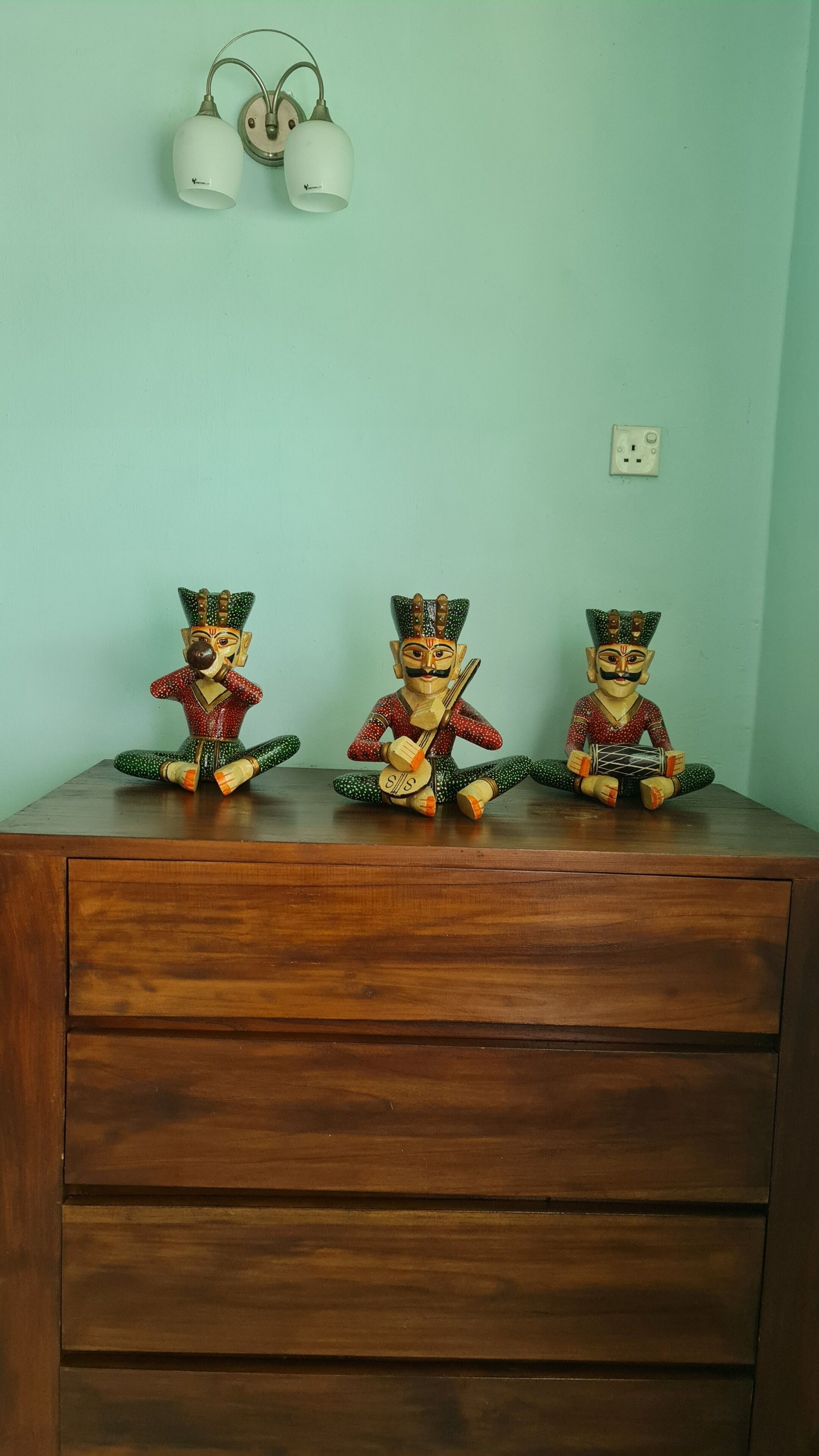 Trois statuettes en bois sont posées sur une commode, devant un mur vert clair. On aperçoit une prise de courant et deux lampes fixées au mur au-dessus.