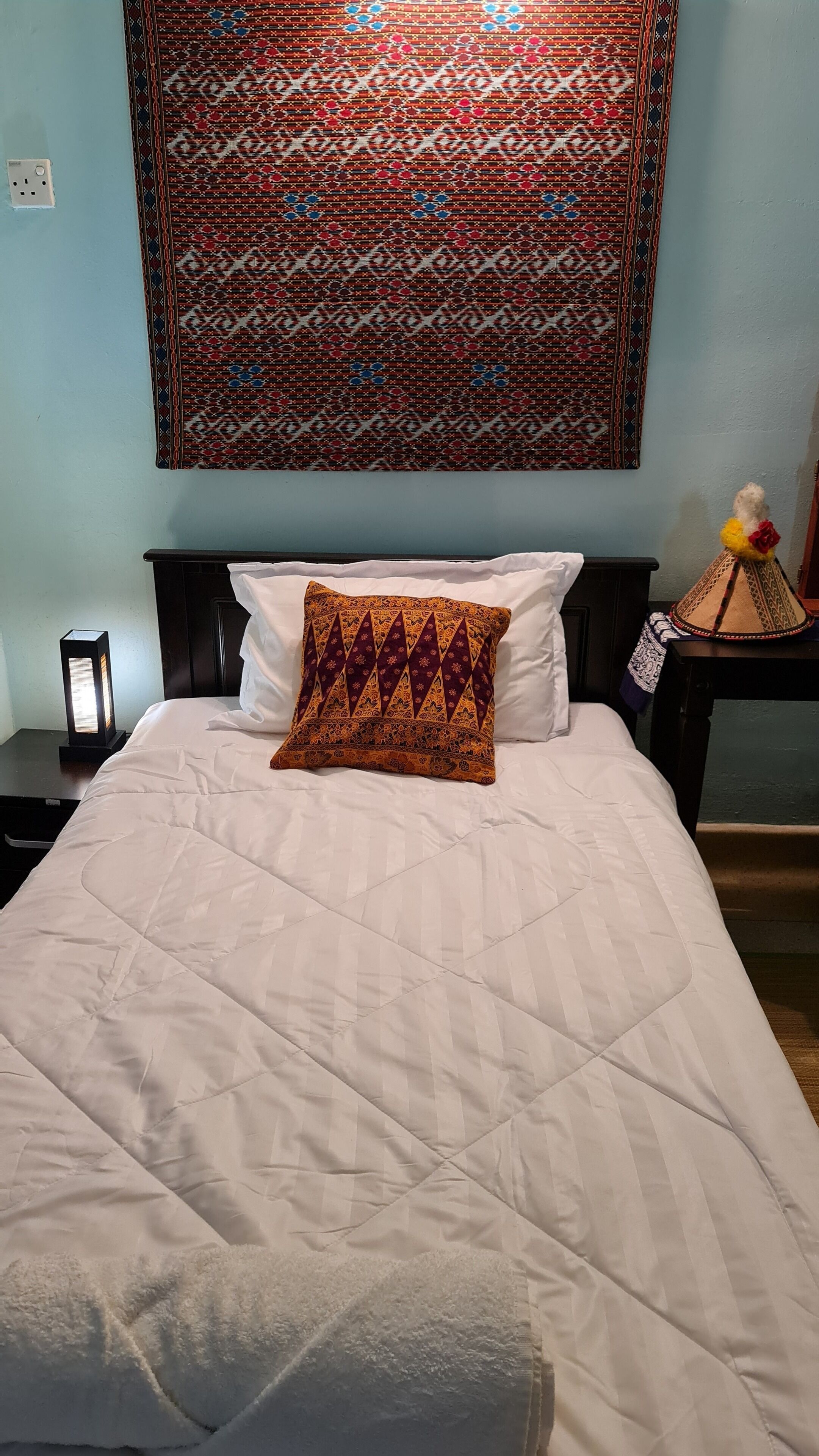 A bed with a white quilt and a patterned pillow, a wall tapestry, and a nightstand with a lamp.