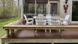 Eine Holzterrasse mit überdachter Veranda, weißen Adirondack-Stühlen und einem Tisch.