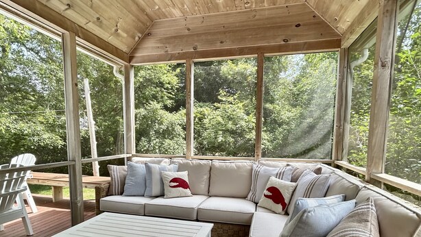 Ein überdachter Außenbereich mit Holzboden, einem weißen Sofa mit gemusterten Kissen und einem Streifen Teppich.