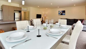 A modern dining area with a long table set for six, white chairs, and a kitchen in the background. - Room to spread out · fenced lawn, pets welcome - Weston Villa (St Albans)
