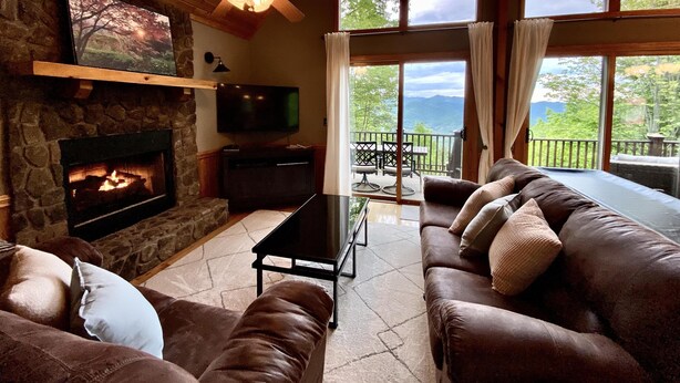 A cozy living room with a stone fireplace, leather sofas, a flat-screen TV, and a large window offering a scenic view. - Mountain Time | Scenic Views, Hot Tub, Wi-Fi, & Shuffleboard (Bryson City)