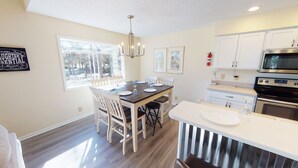 A modern kitchen with white cabinetry, a dining area with a wooden table and chairs, and a large window providing natural light. - Golf Course Comfort Awaits with views (CV4A) (Sunset Beach)