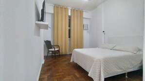 Une petite pièce propre, avec un lit, une chaise, un téléviseur posé sur une étagère et une fenêtre avec des rideaux.