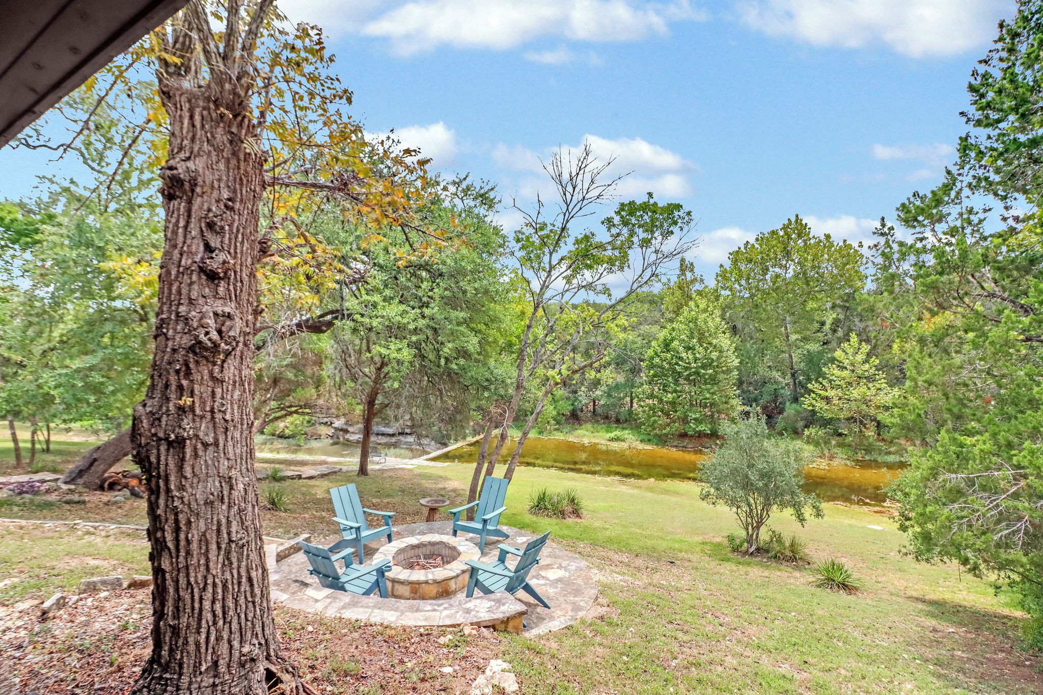 An outdoor seating area with a fire pit and chairs, surrounded by trees and a pond.