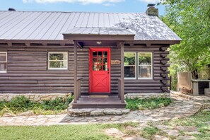 A log cabin with a red door, stone steps, and a clear water log home sign. - Hill Country Log Cabin Near Wimberley | Hot Tub, Fire Pit & Scenic Views (Wimberley)