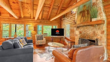 Interior de una cabaña de troncos con televisor, mobiliario de cuero, chimenea de piedra y ventanas amplias.
