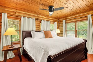A wooden cabin bedroom with a bed, two bedside lamps, a ceiling fan, and large windows.