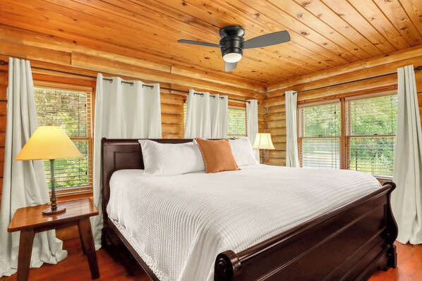 Une chambre dans une cabane en bois, avec un lit, deux lampes de chevet, un ventilateur de plafond et de grandes fenêtres.