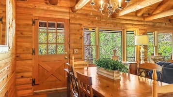 A wooden cabin interior with a dining table, chairs, a chandelier, and large windows.
