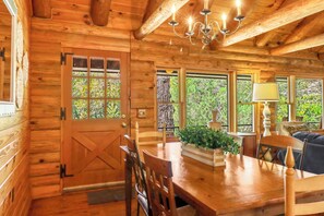 Interior de una cabaña de madera con una mesa de comedor, sillas, un candelabro y ventanas amplias.