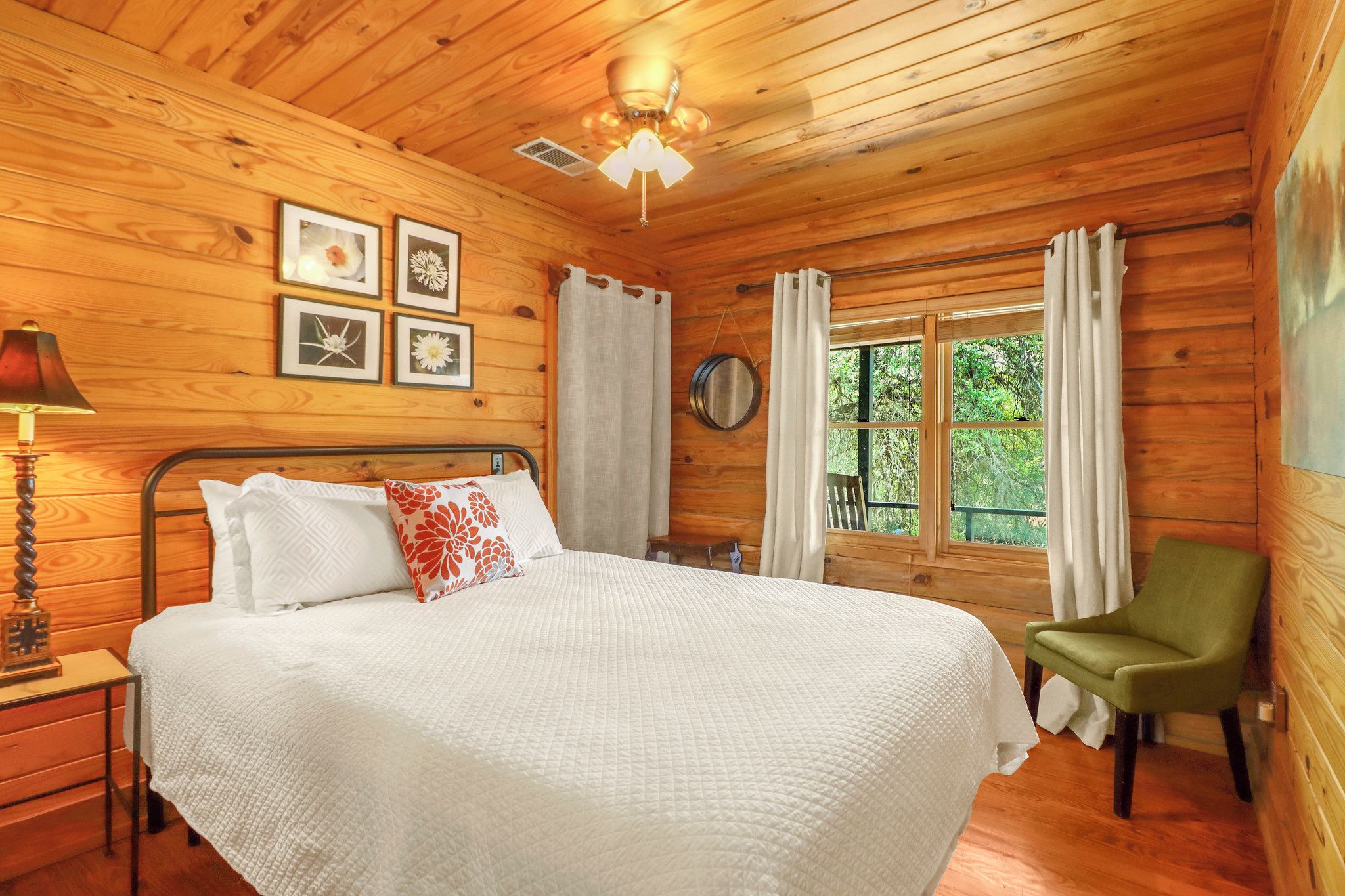 A wooden cabin bedroom with a bed, a nightstand, a lamp, a chair, a window with curtains, and framed pictures on the wall.