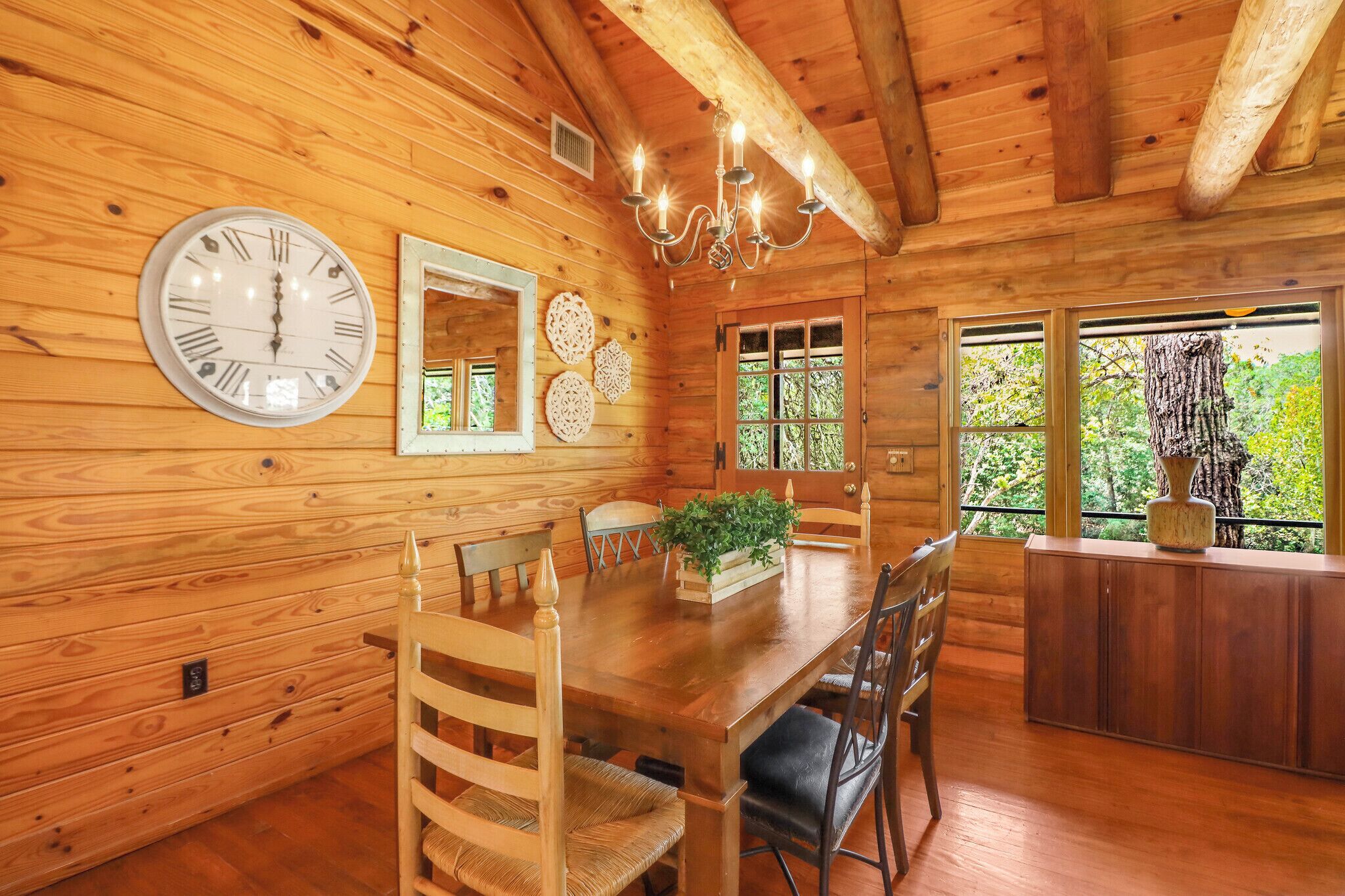 A wooden cabin interior with a dining table, chairs, a clock, decorative wall pieces, and a view through the window.