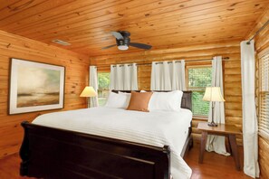 Un dormitorio de cabaña de madera con cama, dos lámparas de noche, un ventilador de techo y una ventana con cortinas.