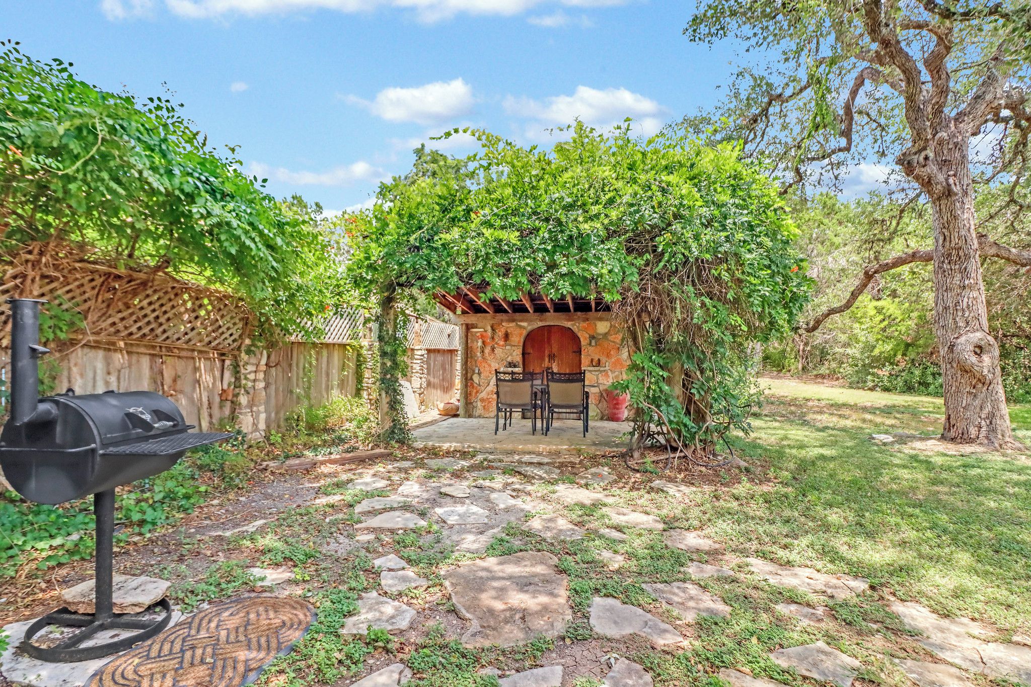 Un jardin avec une terrasse en pierre, un barbecue, une clôture en bois et une arche en pierre menant à un espace de détente abrité.