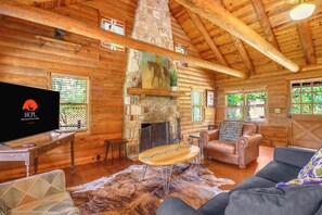 A log cabin interior with a stone fireplace, wooden furniture, and a television. - Hill Country Log Cabin Near Wimberley | Hot Tub, Fire Pit & Scenic Views (Wimberley)