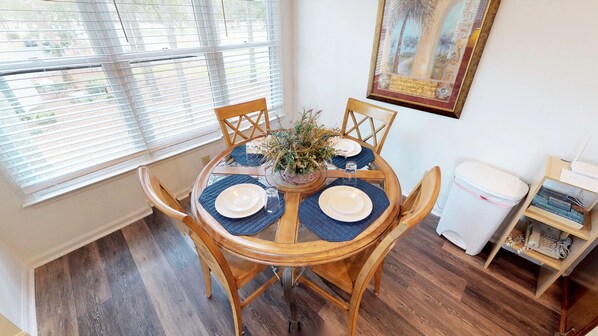 A dining area with a round wooden table set for four, surrounded by wooden chairs. A potted plant is placed in the center of the table.