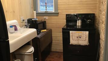 A compact kitchen with wooden walls, a window with a candle, a microwave, a stove, and a sink.