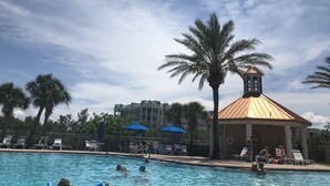 A swimming pool with lounge chairs and umbrellas, surrounded by palm trees and a building with a distinctive roof. - Harbour Village: golf, two beds, two baths, marina, and most spectacular beach.  (Ponce Inlet)