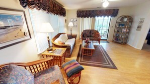 A cozy living room with wicker furniture, a patterned rug, and a view of the outdoors through large windows. - Unwind & Play | River Creek Golf Condo in NC! | RC401 (Sunset Beach)