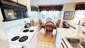 A compact kitchen with a stove, microwave, toaster, and dining area with wicker chairs. - Unwind & Play | River Creek Golf Condo in NC! | RC401 (Sunset Beach)