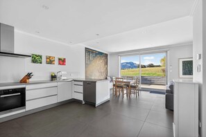 A modern kitchen with a central island, a dining area with a view, and a large window. - Tucker Beach Private, Exclusive Retreat for 1-4 people | Iconic Views | Spa pool (Queenstown)