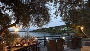 An outdoor dining area with stone flooring, wicker furniture, and a view of a lake and hills in the background. - Beach Front, Romantic Escape, Breathtaking Views, 'Pieds dans L'eau', & More (Magnesia)
