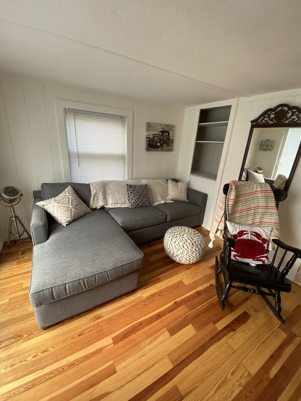 A living room with a grey sectional sofa, a wooden floor, a black rocking chair, and a window with blinds.