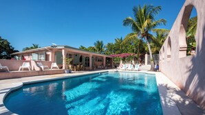 Una piscina con sillones de descanso y una zona de estar cubierta, rodeada de palmeras y un edificio con fachada rosa.