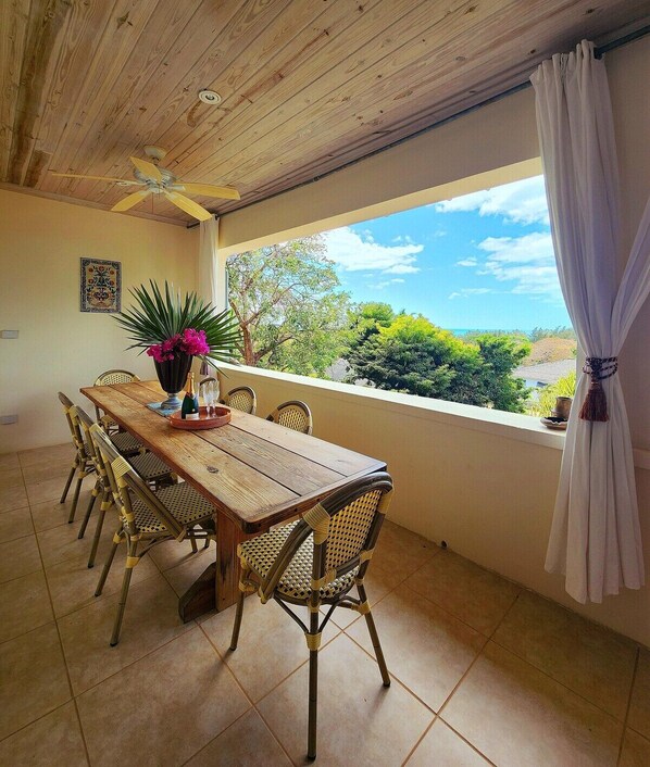 Un comedor con mesa de madera y sillas de mimbre, una planta en maceta y vistas al exterior.
