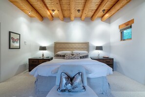 Un dormitorio con techo de madera, una cama grande con muchas almohadas, lámparas de noche y una obra de arte con mariposas enmarcada en la pared.